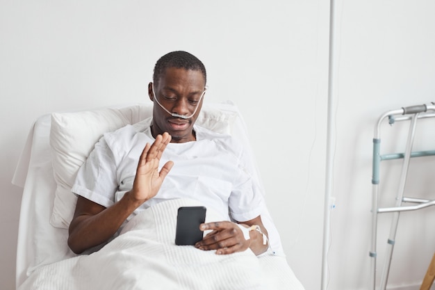 Portret van een Afro-Amerikaanse man die via een videogesprek in het ziekenhuis spreekt en naar de camera zwaait terwijl hij in een wit bed ligt, kopieer ruimte