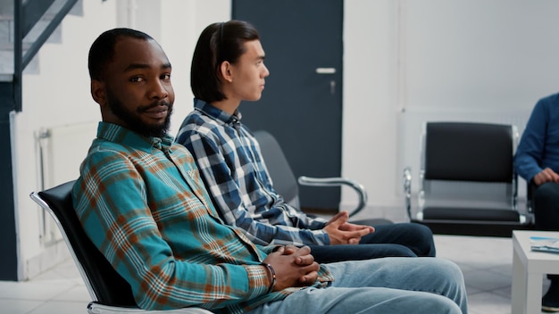 Portret van een Afro-Amerikaanse patiënt zittend op een stoel in de wachtkamer in een medische faciliteit, wachtend om een consultatieafspraak te starten bij de receptie van het ziekenhuis. Diverse mensen in klinische lobby.