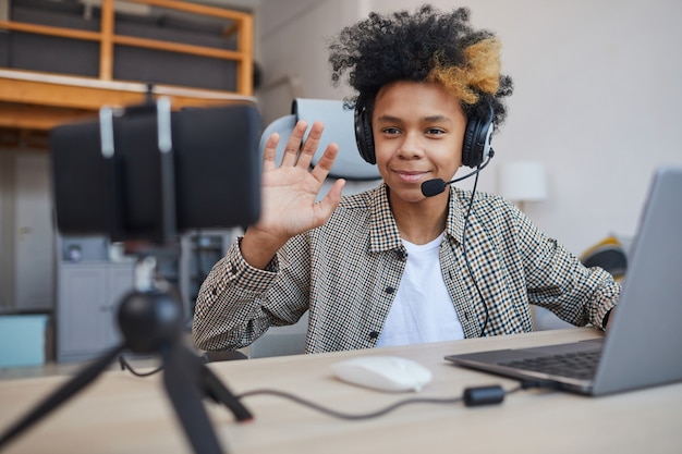 Portret van een Afro-Amerikaanse tienerjongen die een headset draagt en naar de camera zwaait terwijl hij thuis videogames streamt, concept voor jonge gamers of bloggers, kopieer ruimte