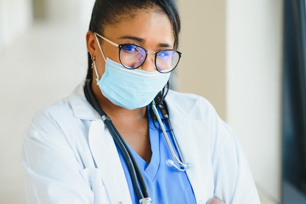 Portret van een Afro-Amerikaanse vrouwelijke arts die een masker draagt dat zich in de ziekenhuisgang bevindt. geneeskunde, gezondheid en gezondheidszorg tijdens de pandemie van het coronavirus covid 19.