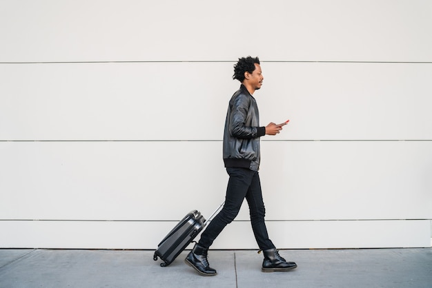 Portret van een afro-toerist die zijn mobiele telefoon gebruikt en een koffer draagt terwijl hij buiten op straat loopt