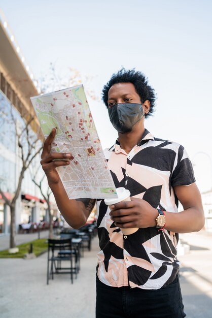 Portret van een afro toeristische man met een beschermend masker en op zoek naar een routebeschrijving op de kaart terwijl hij buiten op straat loopt. Toerisme concept.