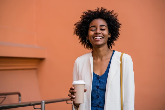 Portret van een afro zakenvrouw glimlachend en met een kopje koffie terwijl ze buiten op straat staat