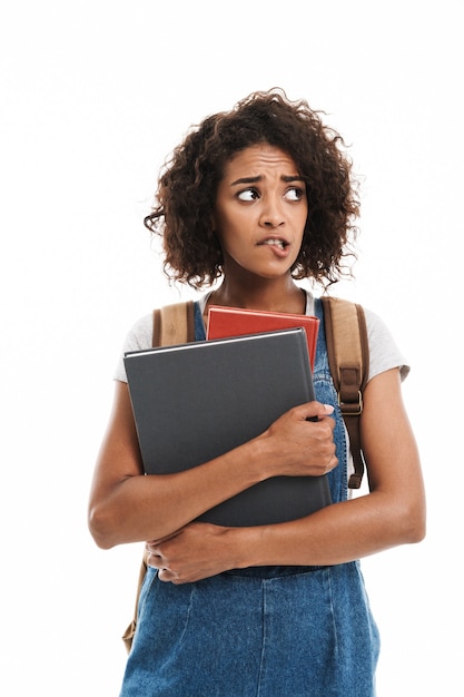 Portret van een angstige vrouw met een rugzak die fronst en oefenboeken vasthoudt die over een witte muur zijn geïsoleerd