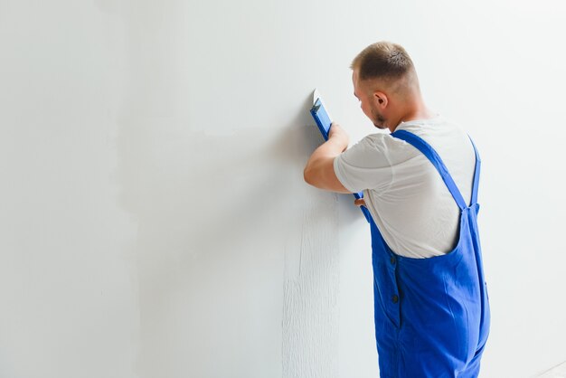 Portret van een arbeider in overall en met een plamuurmes in zijn handen tegen de gepleisterde muurachtergrond. reparatiewerk en bouwconcept