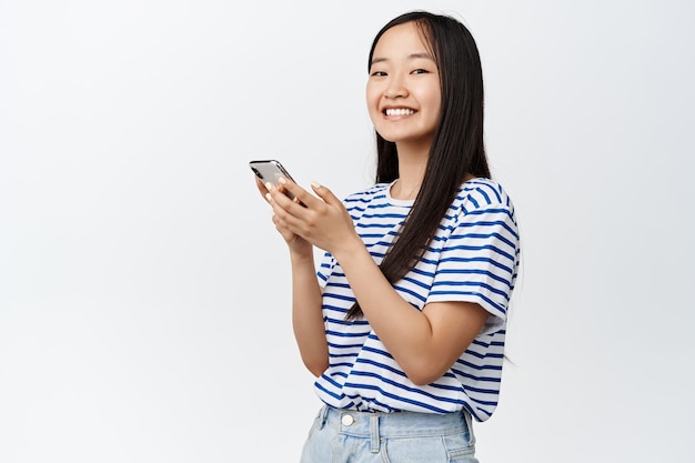 Portret van een Aziatisch meisje dat smartphone gebruikt en glimlacht, concept van cellulaire technologie en mobiele applicaties op wit