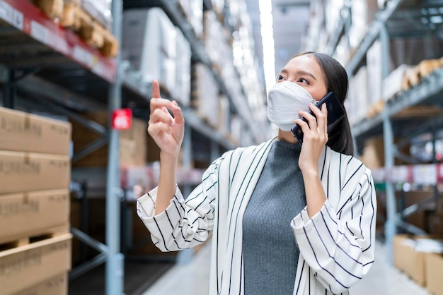 Portret van een aziatische bedrijfseigenaar die een masker draagt, een tablet gebruikt die de hoeveelheid voorraadproductvoorraad op de plank controleert bij een distributiemagazijn, een fabriekslogistieke verzend- en bezorgservice