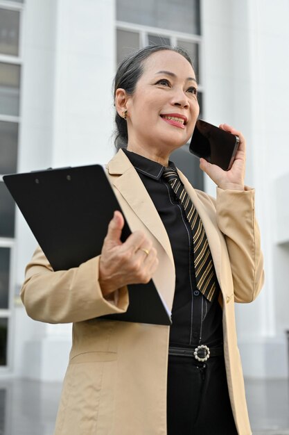 Portret van een Aziatische oude zakenvrouw die een zakelijke telefoongesprek voert aan de buitenkant van het gebouw