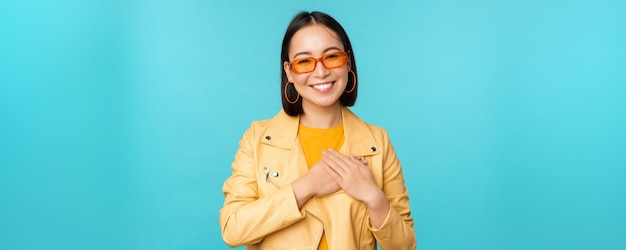 Portret van een aziatische vrouw die lacht, hand in hand op het hart en met tederheid kijkt naar camera dankbare emotie die over blauwe achtergrond staat