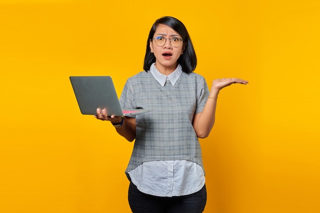 Portret van een Aziatische vrouw met een bril die verrast is met een laptop met een verward gezicht over een gele achtergrond