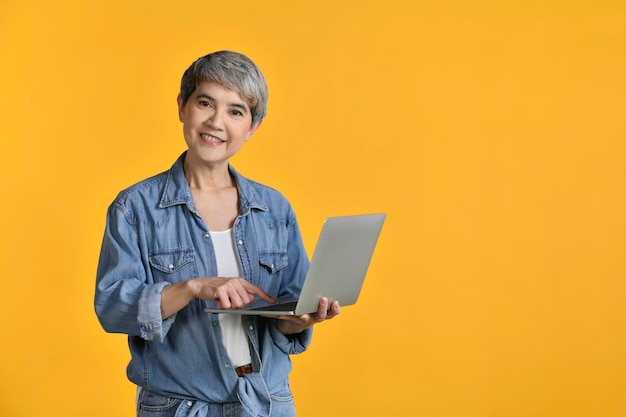 Portret van een Aziatische vrouw van middelbare leeftijd 50s met een casual denim overhemd wit t-shirt met een laptopcomputer en wijzende vingers geïsoleerd op een achtergrond in kleur die naar de camera kijkt en glimlacht
