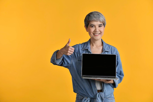 Portret van een Aziatische vrouw van middelbare leeftijd in de vijftig die een laptopcomputer vasthoudt over een achtergrond in kleur