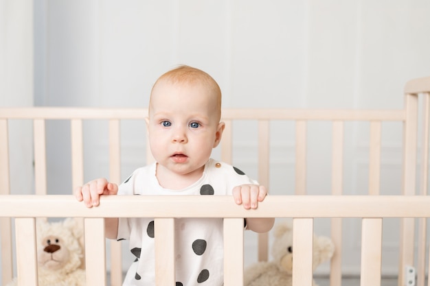 Portret van een baby van 8 maanden oud staande in een wieg met speelgoed in pyjama in een lichte kinderkamer na het slapen en kijken naar de camera, een plek voor tekst