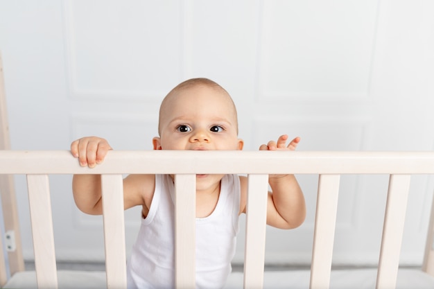 Portret van een babyjongen 8 maanden oud staande in een wieg in een kinderkamer in witte kleren en knabbelen op het bed, baby's ochtend, kinderartikelen concept