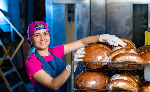 Portret van een bakkersmeisje tegen de achtergrond van planken met vers brood in een bakkerij. Industriële broodproductie