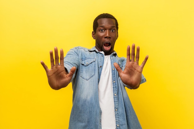 Portret van een bange hysterische jongeman in een casual denim shirt met open mond en opgeheven handen om te verdedigen tegen angst, geschokte en geschokte uitdrukking. studio-opname geïsoleerd op geel