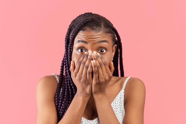 Portret van een bange jonge Afro-Amerikaanse dame die haar gezicht bedekt met handen over roze studio