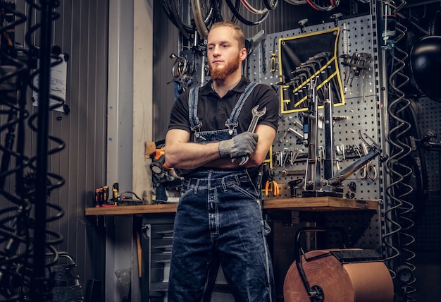 Portret van een bebaarde fietsenmaker met gekruiste armen houdt de bekersleutel vast op de achtergrond van een gereedschapsstandaard in een werkplaats.