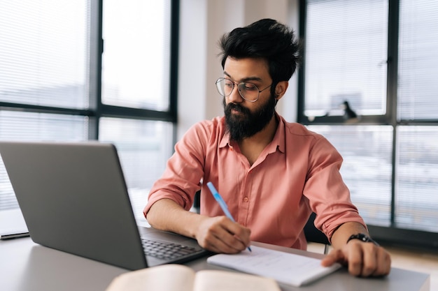 Portret van een bebaarde Indiase zakenman met een bril die een laptop gebruikt en de resultaten van het werkproject analyseert en notities schrijft aan tafel bij het raam