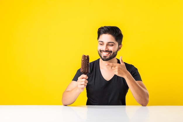 Portret van een bebaarde knappe Indiase jonge man die ijs eet in kegel of ijslolly terwijl hij aan tafel zit tegen een gele studioachtergrond