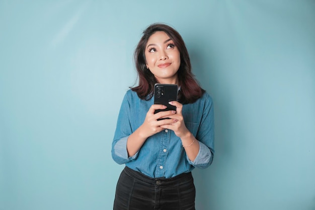 Portret van een bedachtzaam jong casual meisje dat een blauw shirt draagt en opzij kijkt terwijl ze een smartphone vasthoudt die over een blauwe achtergrond wordt geïsoleerd