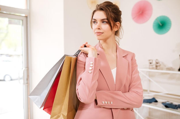 Portret van een bedachtzaam meisje in een roze jas die opzij kijkt met kleurrijke boodschappentassen op haar schouder in de kledingwinkel. Jonge dame staat met zakken in de hand in modeboetiek