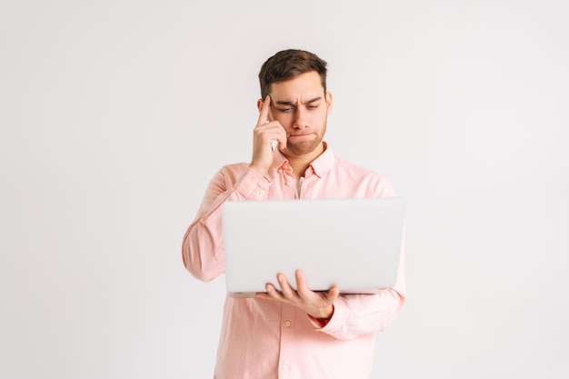 Portret van een bedachtzame jonge man die met een laptopcomputer nieuws leest, video kijkt, op afstand werkt op een witte geïsoleerde achtergrond. Studio-opname van een peinzende man die poseert met een pc die naar het scherm kijkt