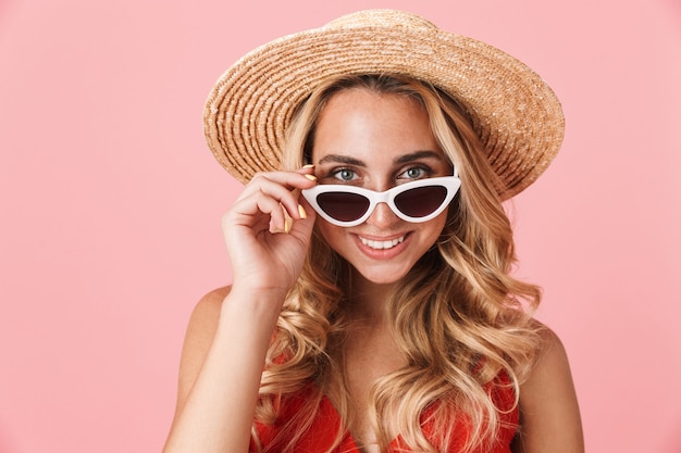 Portret van een behoorlijk opgewonden gelukkige jonge schattige blonde vrouw die zich voordeed over roze muur met hoed en zonnebril.