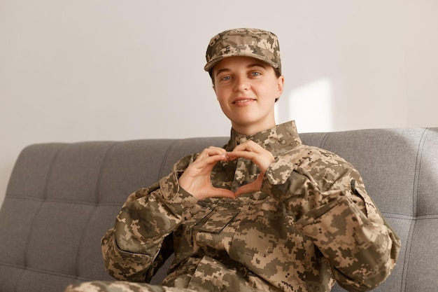 Portret van een blanke glimlachende soldaat die een camouflage-uniform draagt dat op de bank zit en naar de camera kijkt met een positief teken van hartliefde met handen die thuiskomen van het leger