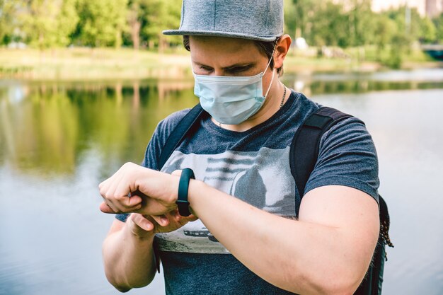 Portret van een blanke man in een medisch masker tijdens een pandemie in de stad maakt gebruik van een smartwatch. nieuw normaal.