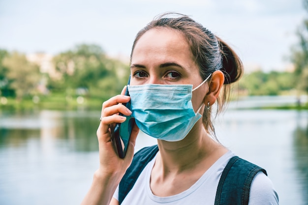 Portret van een blanke meisje in een medisch masker tijdens een pandemie in de stad maakt gebruik van een smartphone. Nieuw normaal.