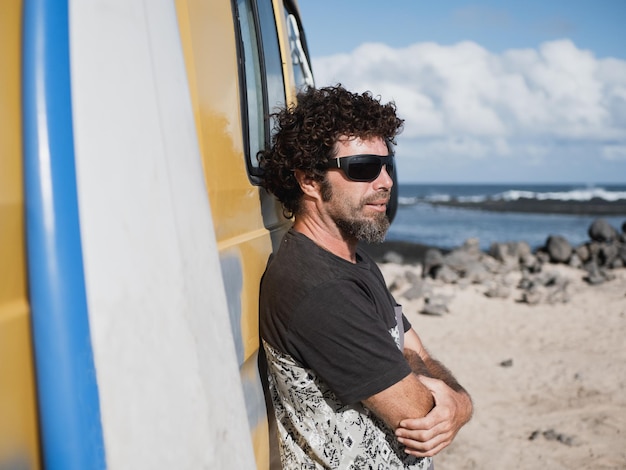 Portret van een blanke nomade aan zee, leunend op zijn camper. Zijn surfplank staat ernaast