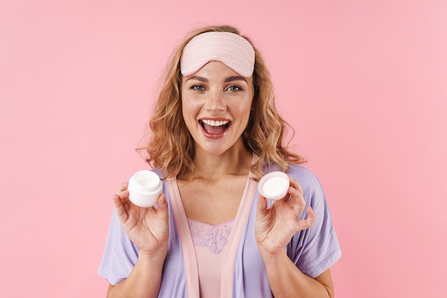 Portret van een blanke vrolijke vrouw met een slaapmasker die lacht terwijl ze gezichtscrème vasthoudt die op roze wordt geïsoleerd