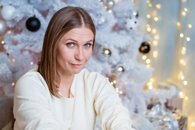 Portret van een blanke vrouw in een lichte, lichte trui die bij de kerstboom zit met een peinzende blik