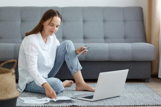 Portret van een blanke vrouw met een wit overhemd en een spijkerbroek zittend op de vloer in de buurt van de bank met behulp van een laptop met een creditcard en een mobiele telefoon die naar een papieren document kijkt