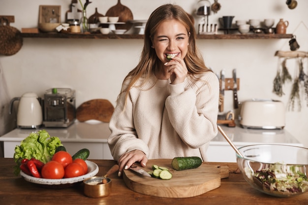 portret van een blije schattige vrouw die vrijetijdskleding draagt en komkommer eet in een gezellige keuken