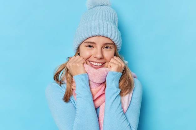 Portret van een blije tedere Europese vrouw draagt een warme wintermuts en sjaal om de nek, voelt gezellig en opgetogen poses tegen een blauwe studioachtergrond en geniet van een goede dag. Mensen seizoen kleding concept