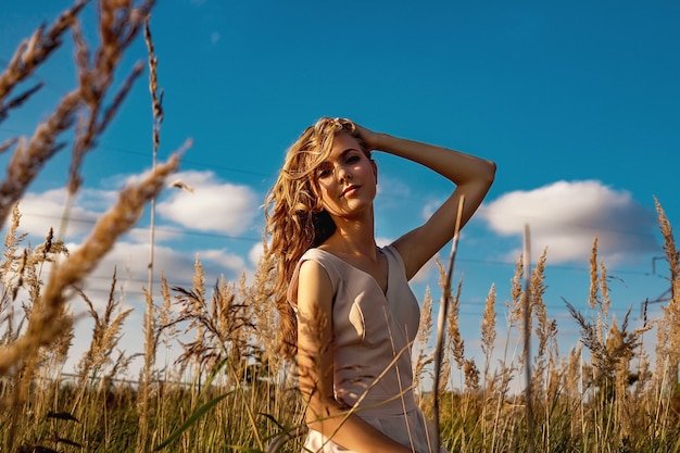 Portret van een blond meisje in een jurk die in een veld met hoog gras en een berk zit.