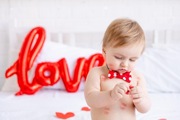 Portret van een blonde babyjongen zittend op een bed in luiers met kussen van rode lippenstift thuis en glimlachend tussen harten en een grote ballon met de inscriptie hou van het concept van Valentijnsdag