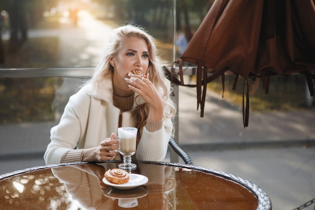 Portret van een blonde dame aan een tafel in een café die een taart, lang haar, een beige jas eet.