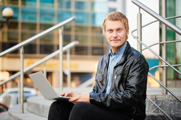 Portret van een blonde man met een baard in een leren jas en shirt, zittend op de trap met de computer op schoot, op de achtergrond het gebouw met een glazen gevel