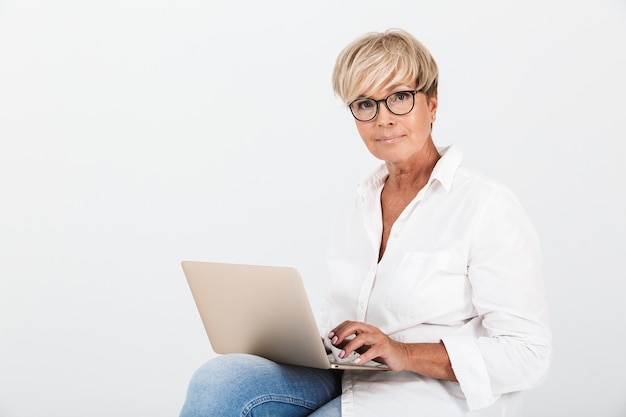 Portret van een blonde volwassen vrouw met een bril die zit met een laptopcomputer die over een witte muur in de studio wordt geïsoleerd