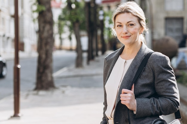 Foto portret van een blonde volwassen zakenvrouw die lacht in een grijze jas op een achtergrond van de stad