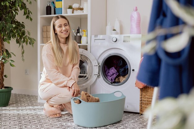 Portret van een blonde vrouw die lacht, gekleed in roze joggingbroek en pantoffels knielt in de wasruimte