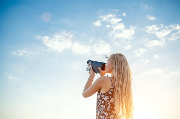 Portret van een blonde vrouw in een jurk met bloemenprint met een vintage videocamera in een druivenveld neemt video op van een opstijgend vliegtuig