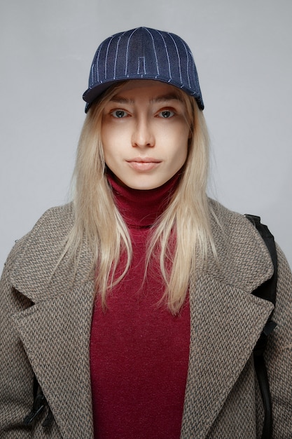 Portret van een blondemeisje in wollen jas en honkbalhoed zonder make-up.