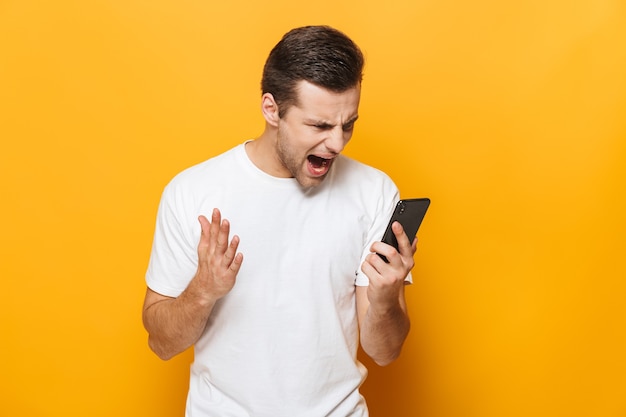 Portret van een boze jonge man met een t-shirt dat geïsoleerd over een gele muur staat, mobiele telefoon gebruikt, schreeuwt