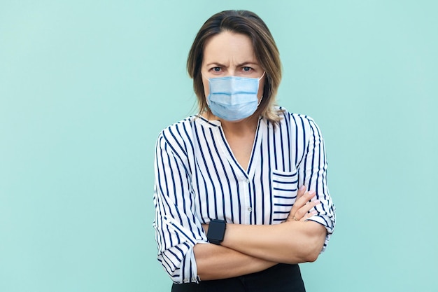 Portret van een boze vrouw van middelbare leeftijd met een chirurgisch medisch masker met gekruiste armen en