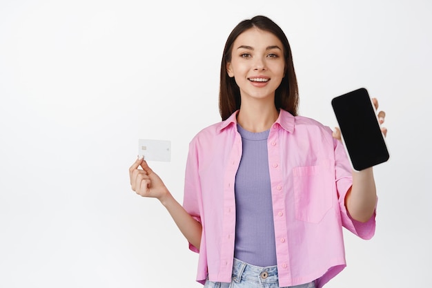 Portret van een brunette vrouw die het scherm van een mobiele telefoon en een creditcard laat glimlachen terwijl ze een toepassing aanbeveelt die op een witte achtergrond staat