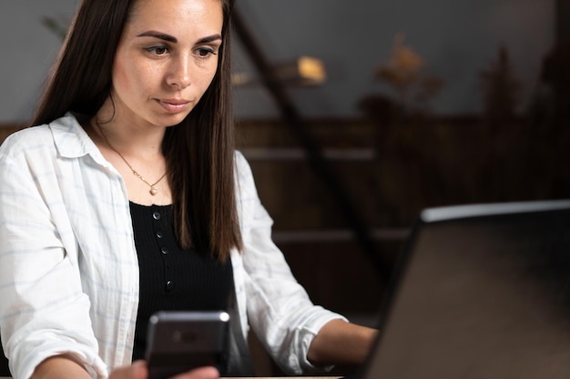 Portret van een brunette vrouw die op kantoor werkt met een laptop die tekst op het toetsenbord typt terwijl ze naar de computer kijkt met een smartphone